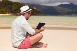 young casual man sitting on the beach and reading on his touchscreen tablet